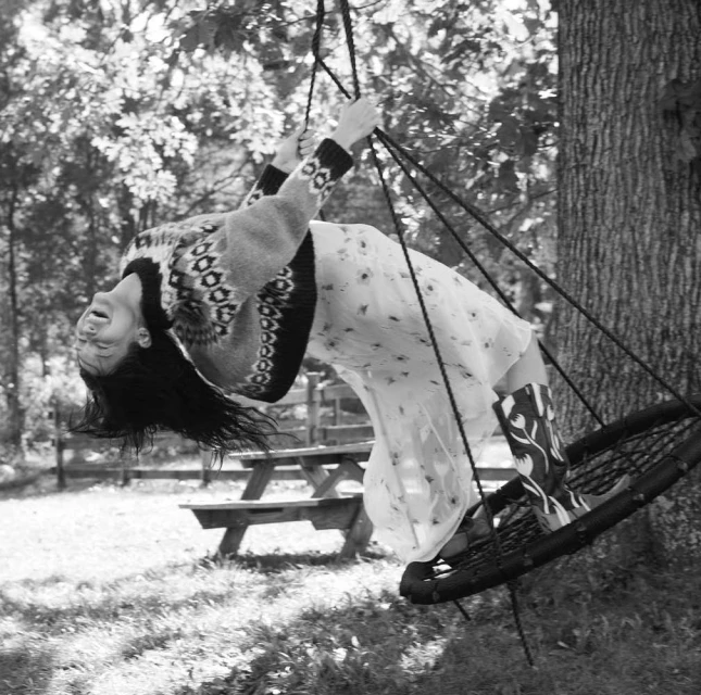Monochrome image of a woman on a swing, laughing with her head tilted back, in a dress, sweater, and boots in an outdoor setting.
