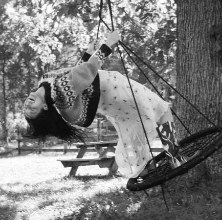 Monochrome image of a woman on a swing, laughing with her head tilted back, in a dress, sweater, and boots in an outdoor setting.