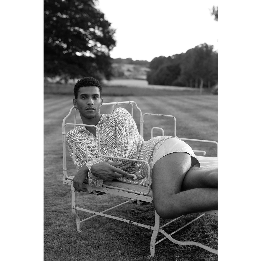 A black and white portrait of a male model lying on a vintage metal lawn chair in a grassy field with trees and sky visible.