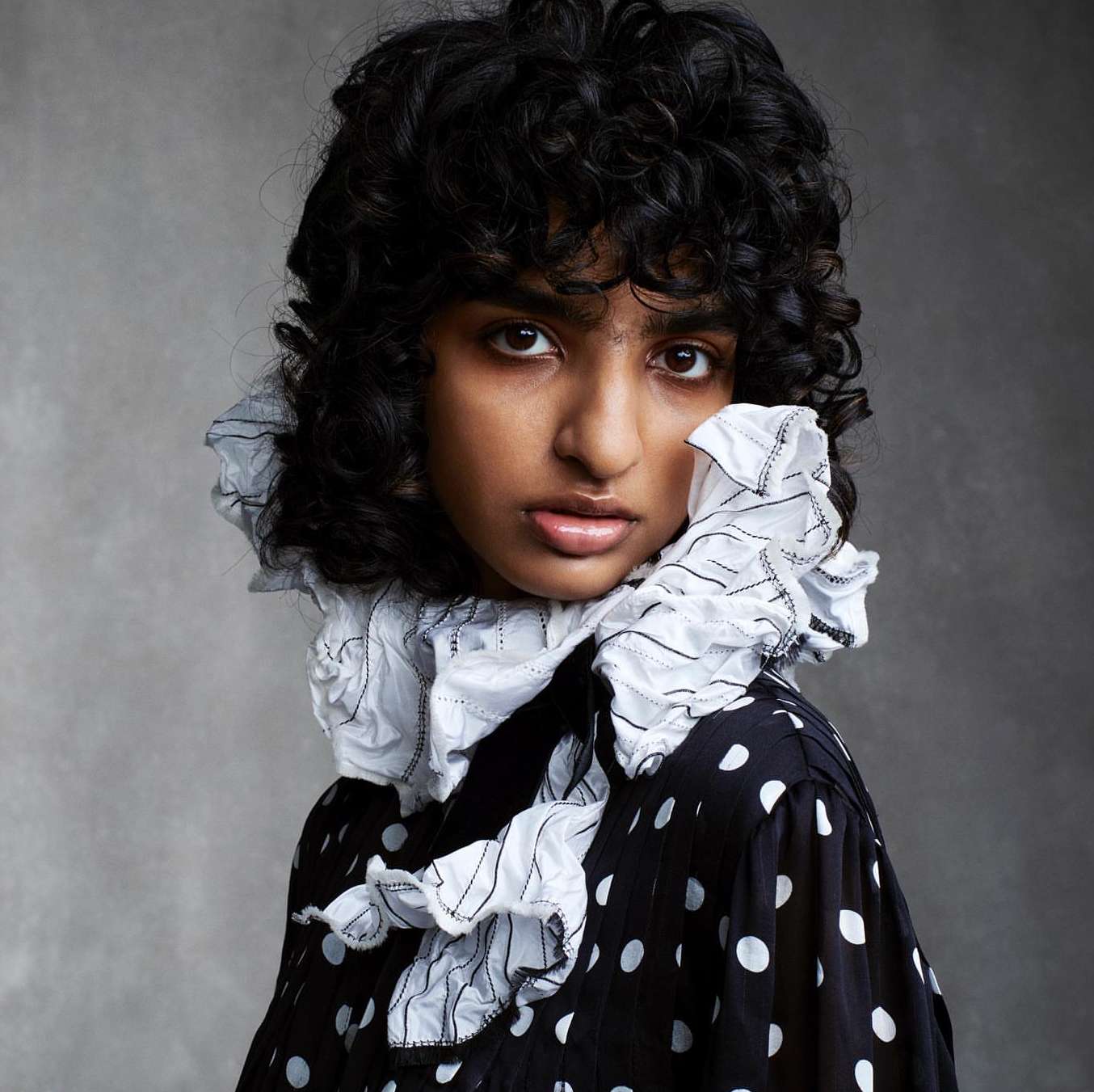 Close-up studio portrait of a young, dark-skinned woman with curly black hair wearing a black and white polka dot dress with a ruffled collar.
