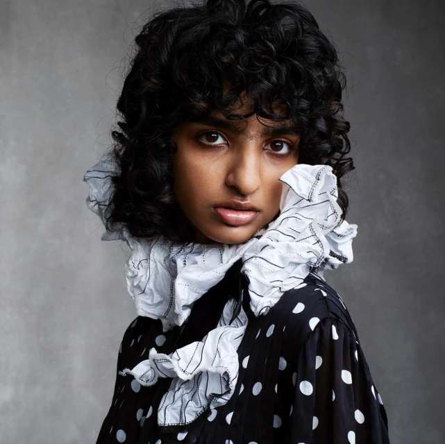 Close-up studio portrait of a young, dark-skinned woman with curly black hair wearing a black and white polka dot dress with a ruffled collar.