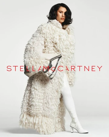 Model in fluffy cream shaggy coat holding a white handbag with white boots against a clean studio background