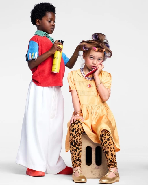 Two children, an African-American boy styling a Caucasian girl's hair with hairspray and curlers against a white backdrop in a studio setting.