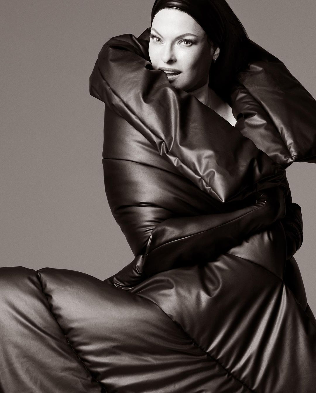 Fashionable woman in oversized puffer coat and head covering poses against a neutral backdrop. Black and white, studio lighting.