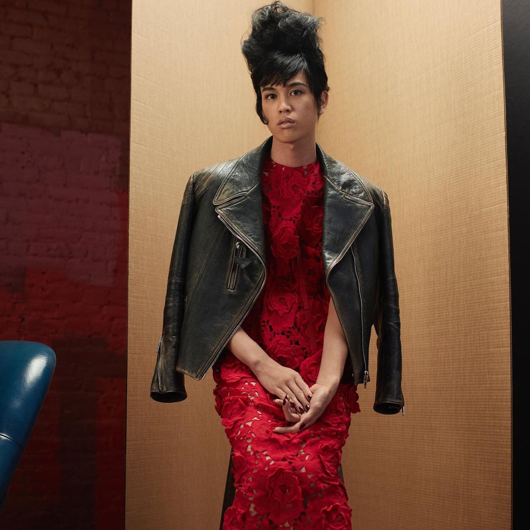 Adult posing in a red floral dress with a leather jacket, standing indoors against a tan and red paneled wall in a medium shot.