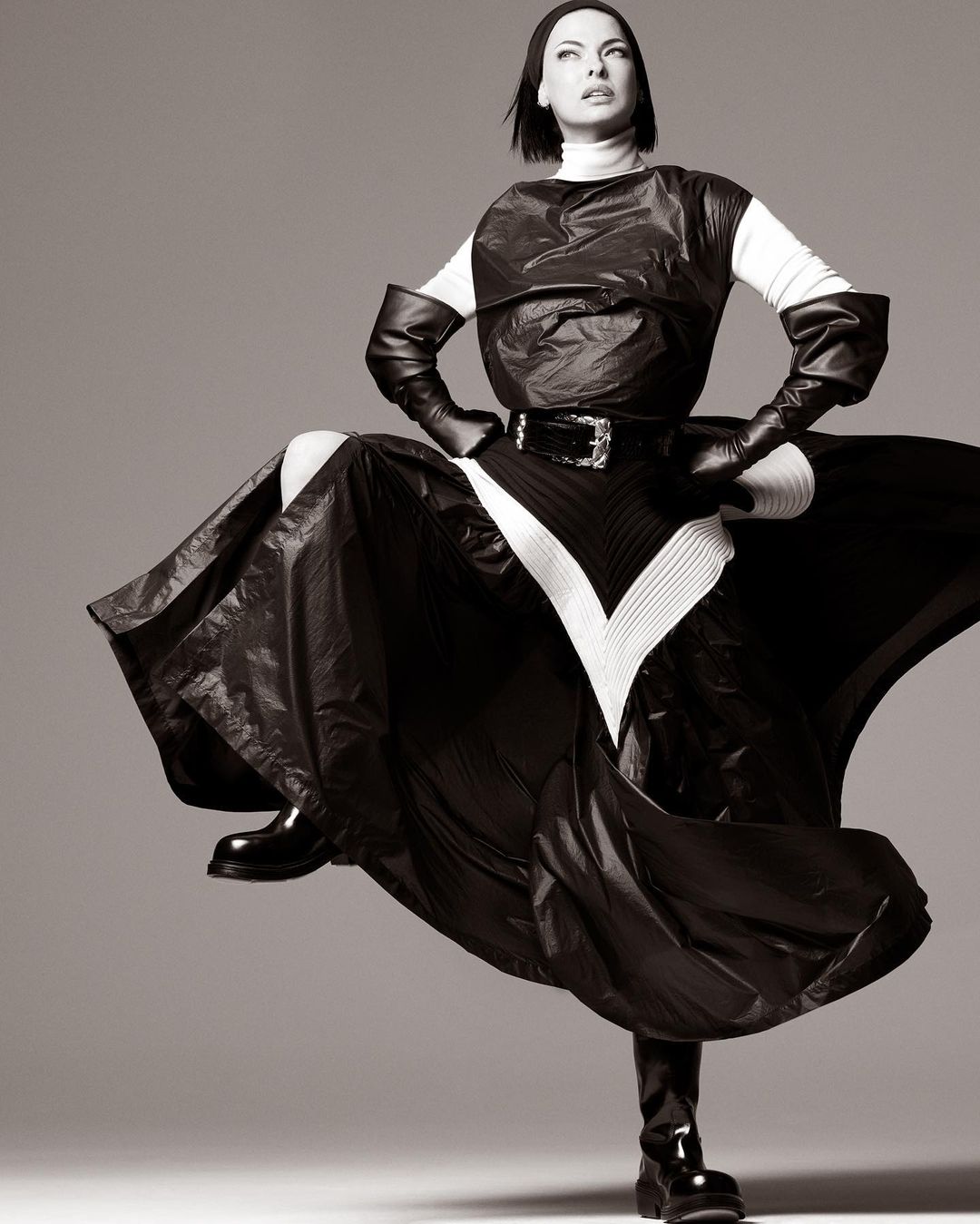 Woman strikes a pose in dramatic black and white studio shot, wearing a high-fashion garment with shiny black fabric and matching boots.