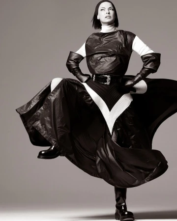 Woman strikes a pose in dramatic black and white studio shot, wearing a high-fashion garment with shiny black fabric and matching boots.
