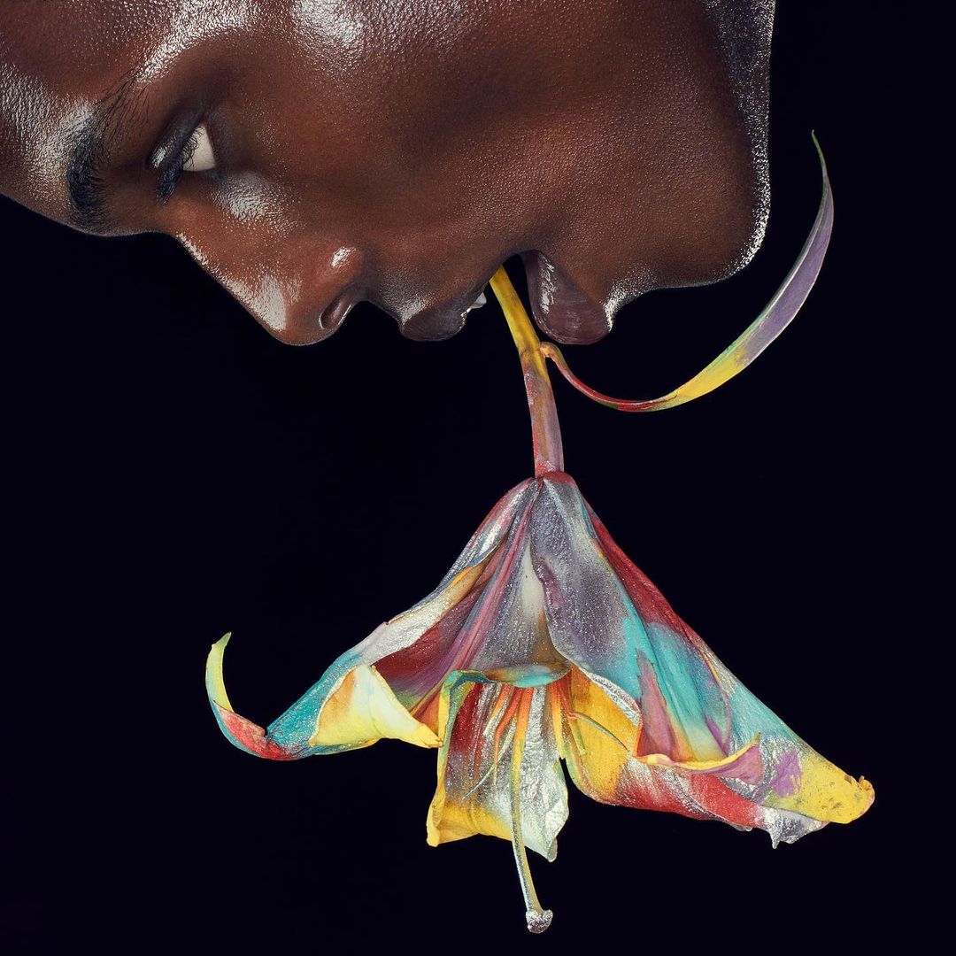 Close-up profile of a black person biting into a vibrant, multi-colored painted lily against a solid black background.