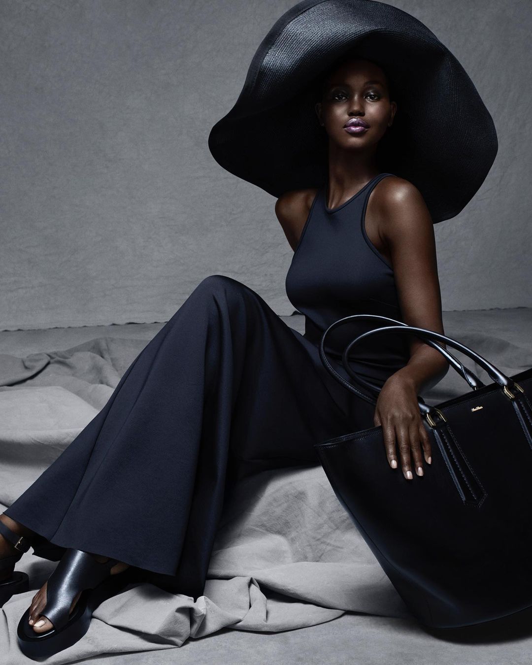 A stylish Black woman sits gracefully in a navy outfit with a wide-brimmed black hat and a large matching tote bag in a studio setting.