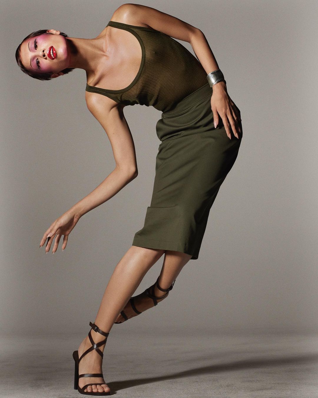 Adult female model in green top and pencil skirt poses with bent pose in studio, wearing strappy sandals and silver bracelet.