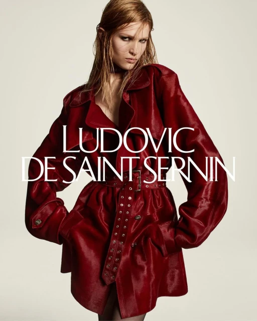 A model in a bold red belted trench coat poses indoors against a neutral backdrop, showcasing a high-fashion editorial look.