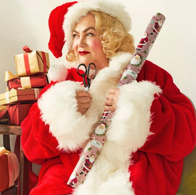 Adult female in Santa Claus costume holds Christmas-themed wrapping paper and scissors against a light-colored indoor backdrop.