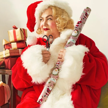 Adult female in Santa Claus costume holds Christmas-themed wrapping paper and scissors against a light-colored indoor backdrop.