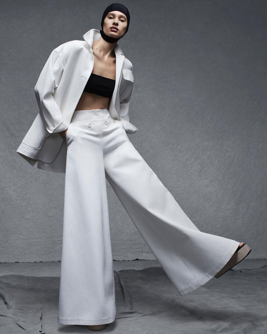 Fashion shot: Woman poses in white wide-leg pants, black top, and white coat with black head covering against a gray backdrop.