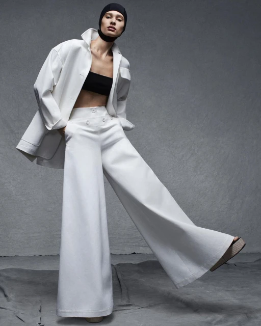 Fashion shot: Woman poses in white wide-leg pants, black top, and white coat with black head covering against a gray backdrop.