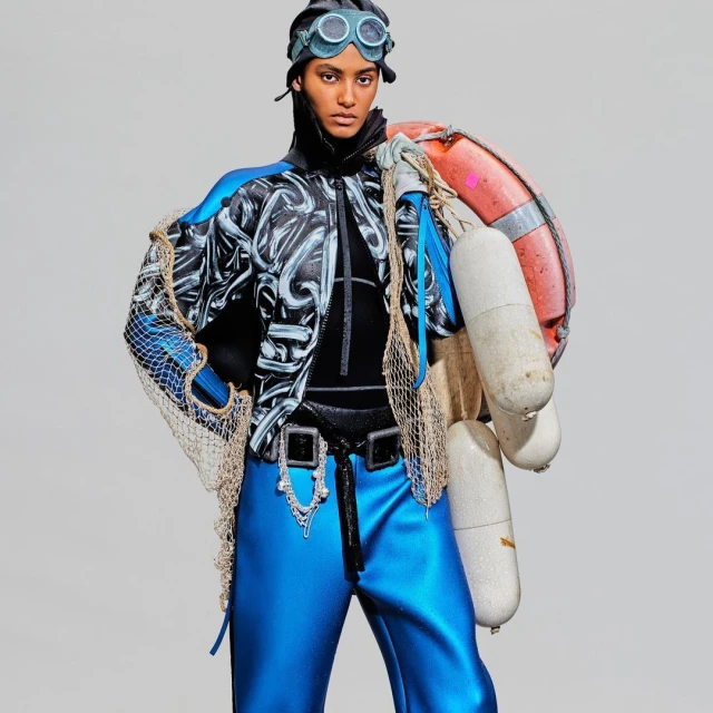 Fashion model in a conceptual blue and black outfit with goggles, carrying life preservers and a fishing net against a white background.