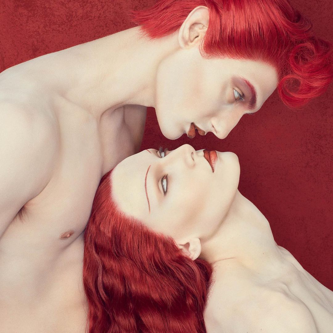 Surreal art featuring a pale-skinned man and woman with bright red hair against a matching background, emphasizing color contrast.