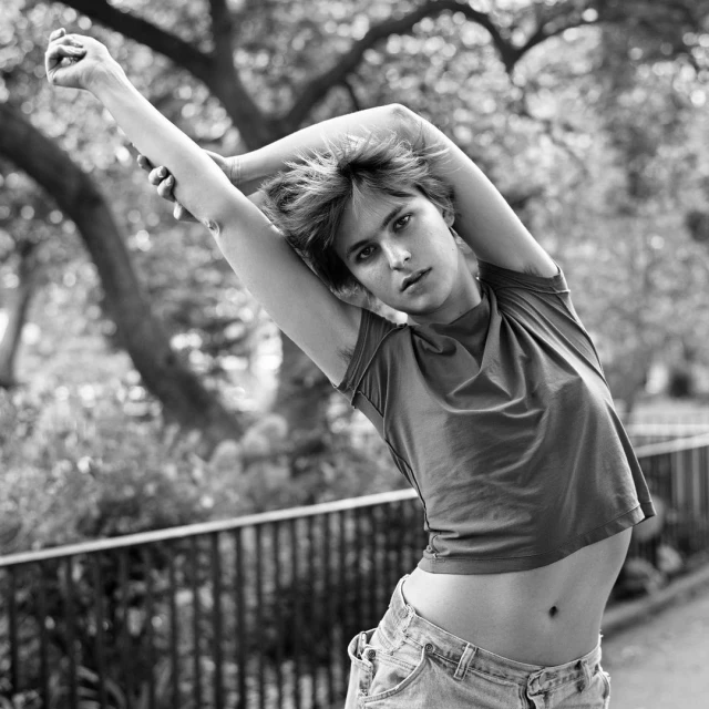 Black and white portrait of a young adult woman with short hair, arms raised above her head, wearing a crop top and jeans, outdoors.