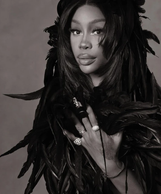 Monochromatic portrait of SZA in feather garment, wearing multiple rings, against a grey backdrop in a studio setting.