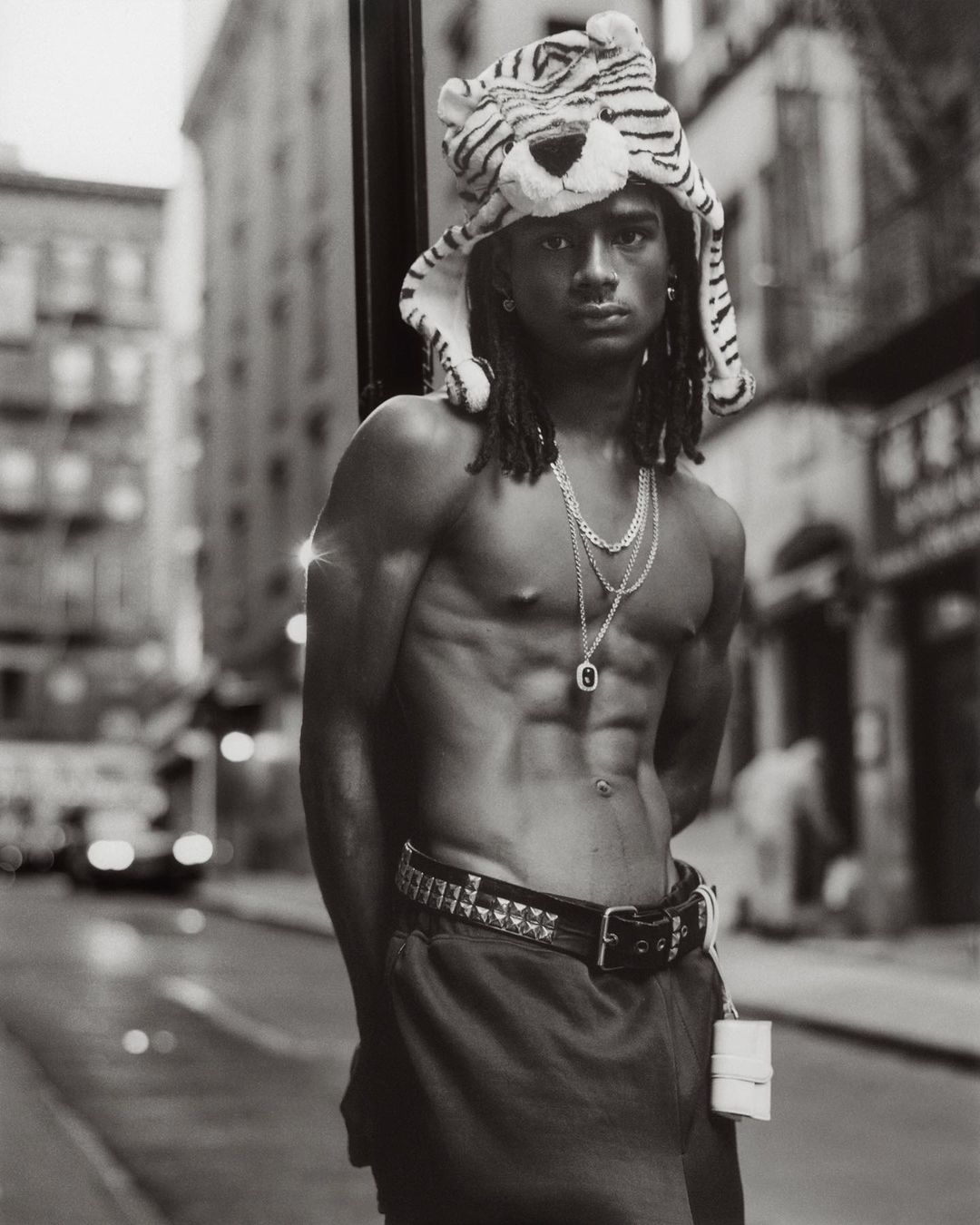 Monochrome portrait of shirtless man with abs wearing a tiger hat, necklaces, and studded belt in a city street setting.