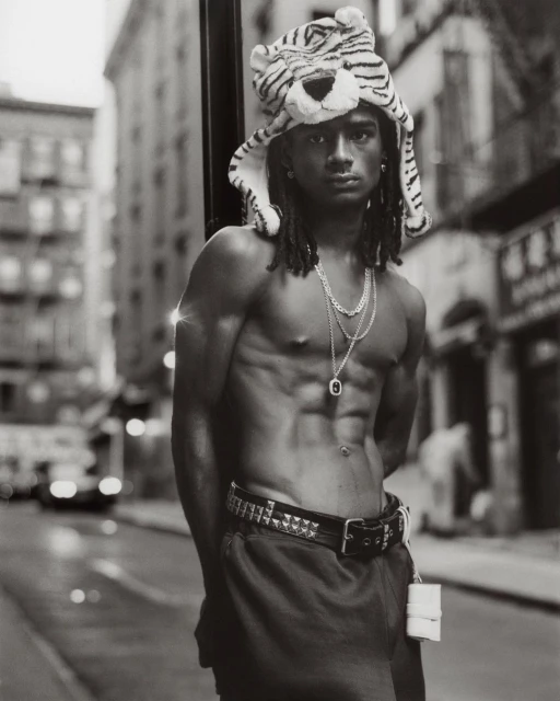 Monochrome portrait of shirtless man with abs wearing a tiger hat, necklaces, and studded belt in a city street setting.
