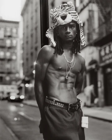 Monochrome portrait of shirtless man with abs wearing a tiger hat, necklaces, and studded belt in a city street setting.