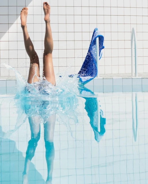 Woman dives into a pool, legs above water, creating splash, with a blue towel and white tiled wall in the background.
