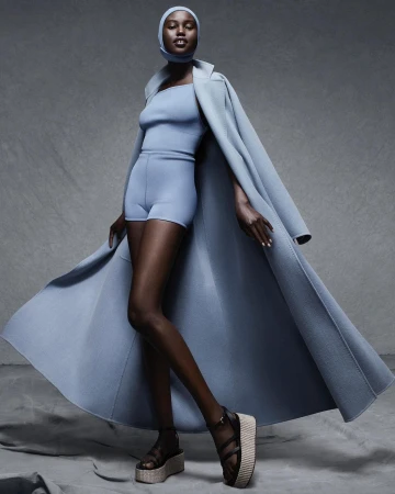 A Black woman is posing in a studio wearing a light blue matching set consisting of a top, shorts, coat, and head covering.