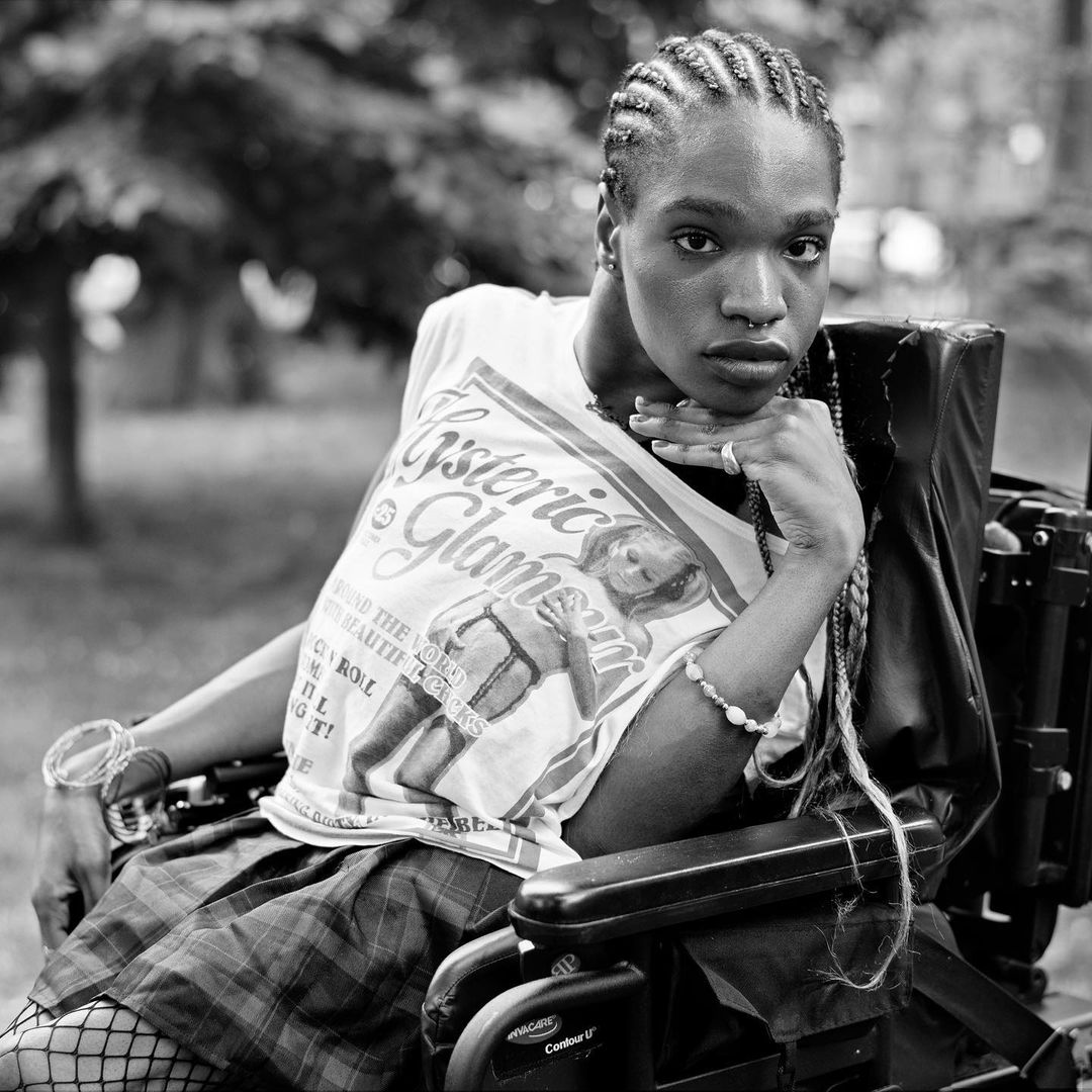 Black and white portrait of a person in a wheelchair, wearing a graphic t-shirt and plaid skirt in a grassy outdoor setting.