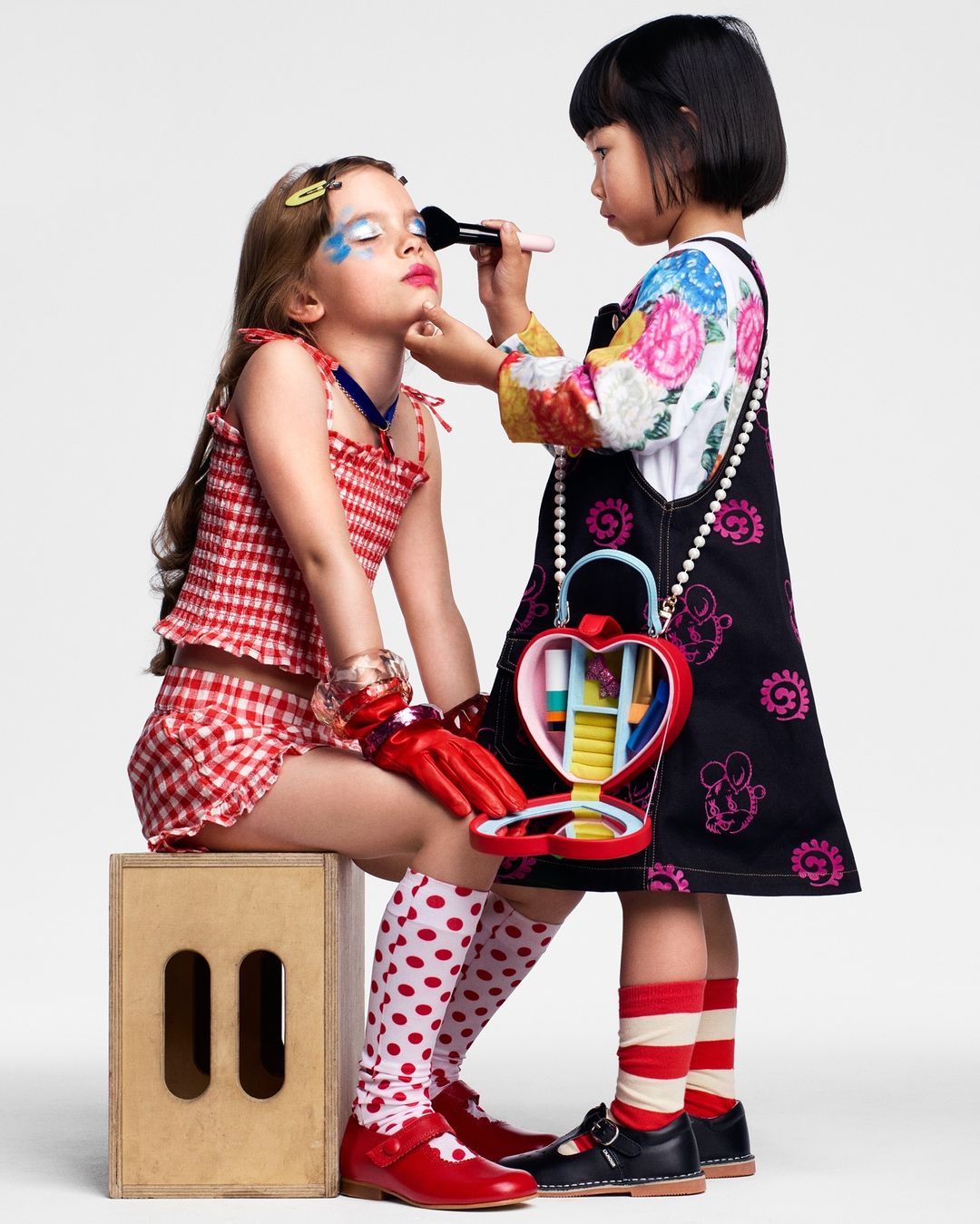 Two young girls play with makeup indoors: one seated with makeup applied, the other applying makeup with a brush. Fun and playful.