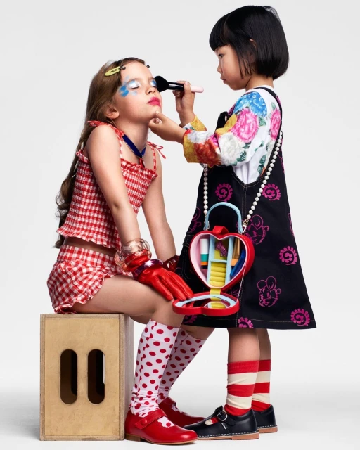 Two young girls play with makeup indoors: one seated with makeup applied, the other applying makeup with a brush. Fun and playful.
