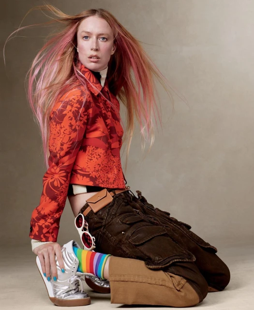 Woman in orange floral jacket and brown cargo pants poses kneeling with silver sneaker, showcasing fashion-forward editorial style.