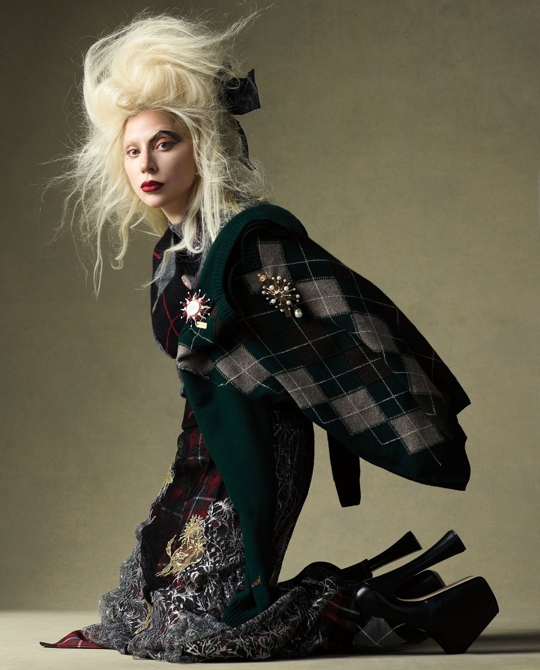 A fair-skinned woman with blonde hair, red lips, and a tartan outfit poses kneeling with stacked platform heels in a studio shot.