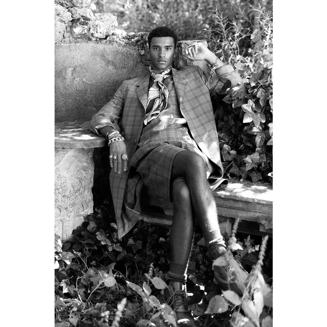 Black and white portrait of a Black male model in a plaid suit sitting outside on a stone bench surrounded by foliage.
