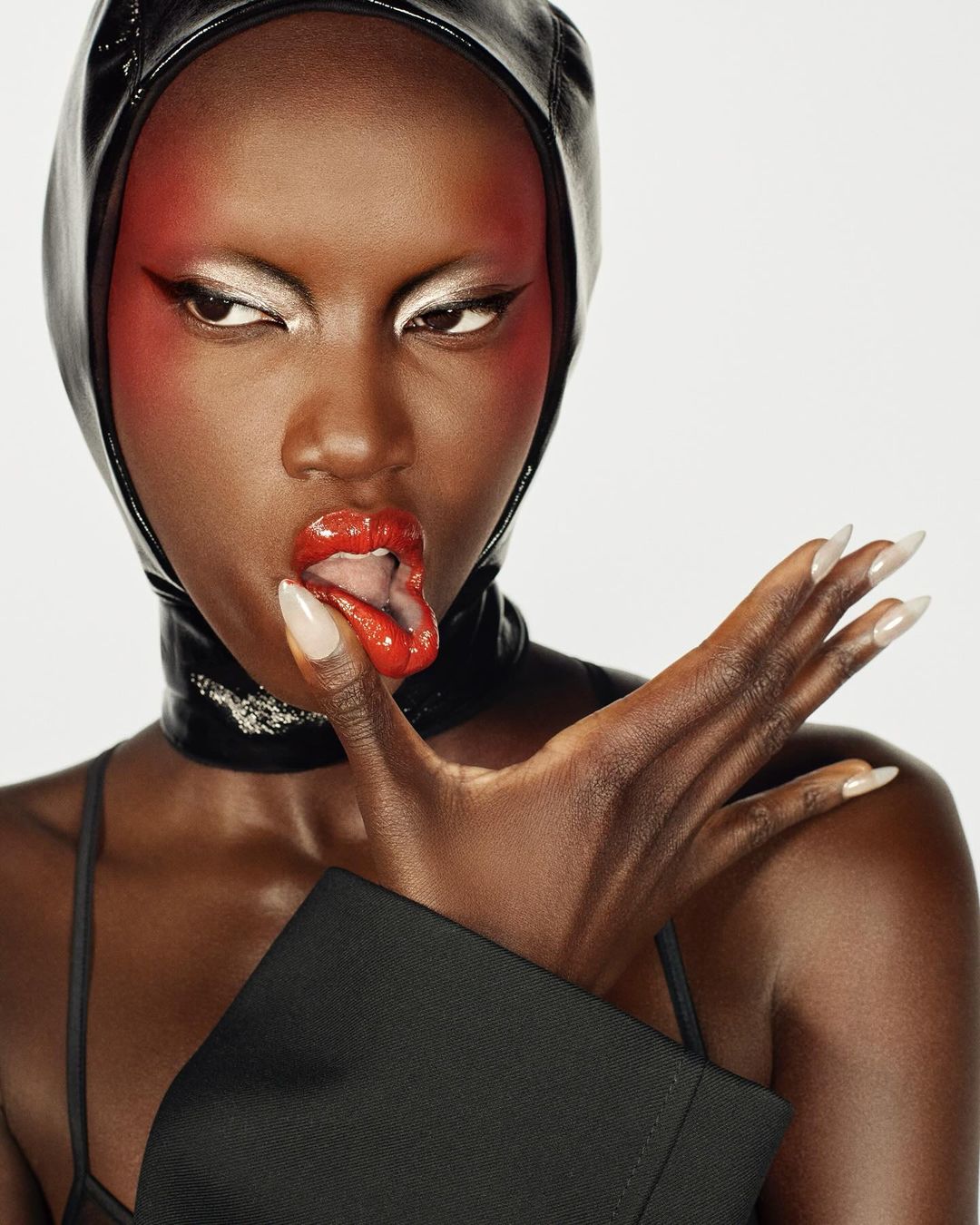 Close-up portrait of a Black woman wearing a black hood and jacket, glossy red lipstick, and silver eyeshadow against a white backdrop.