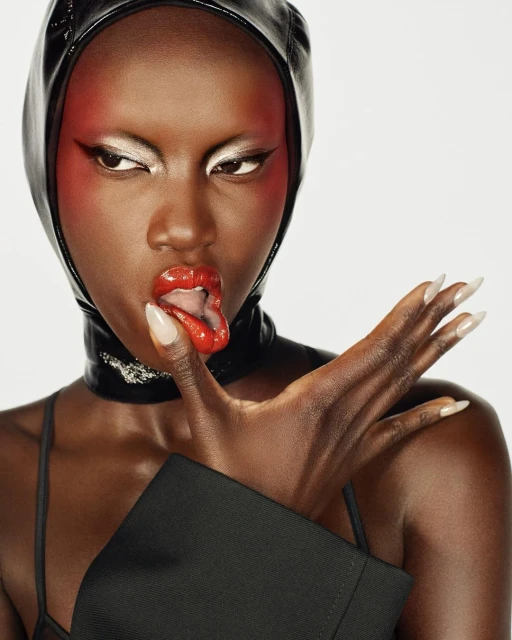 Close-up portrait of a Black woman wearing a black hood and jacket, glossy red lipstick, and silver eyeshadow against a white backdrop.