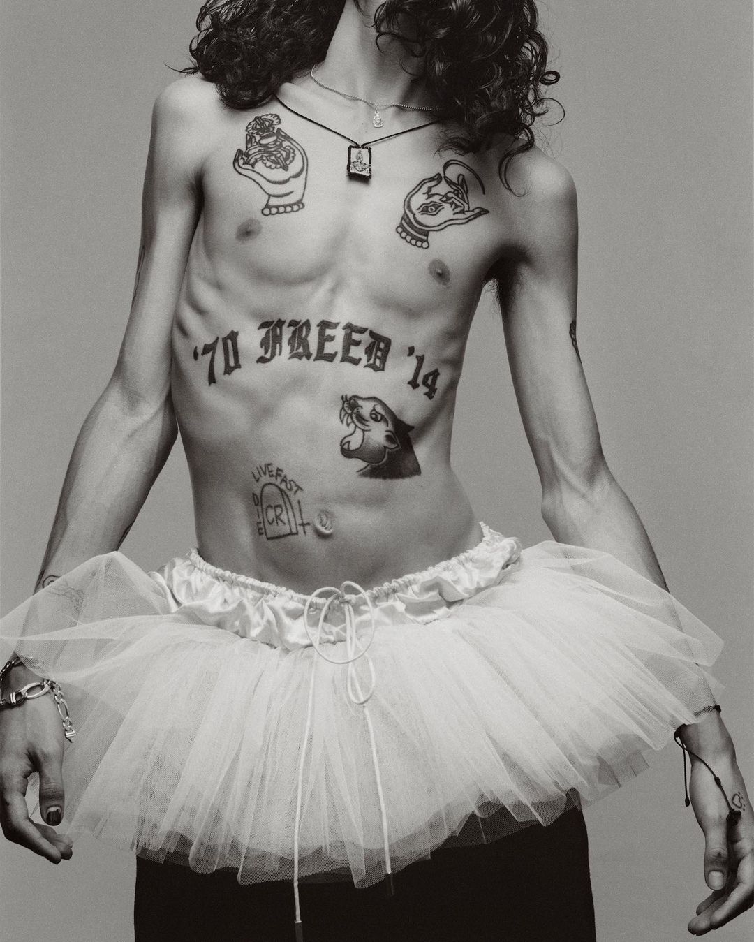 Tattooed male torso with tutu against grey backdrop; expressive editorial fashion portrait showcasing unique style and body art in studio setting.
