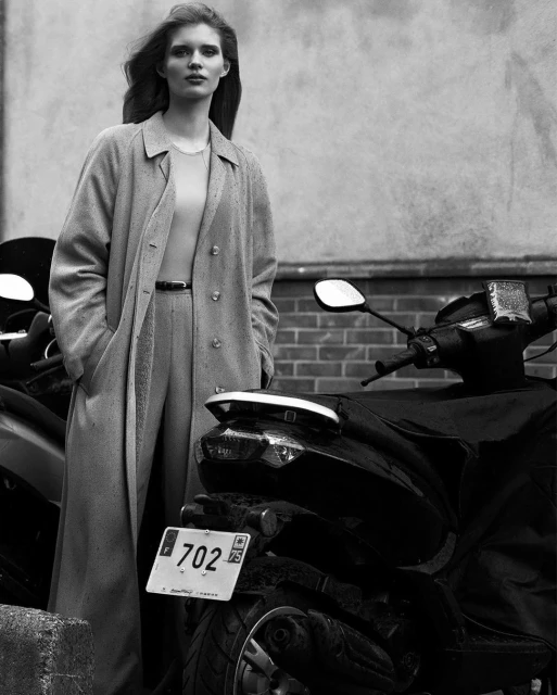 Monochrome portrait of a stylish adult woman in a long coat standing near motor scooters in an urban setting with neutral tones.