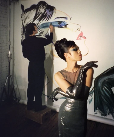 Fashion shoot: A woman in a grey dress and long gloves poses in front of a large painting being worked on by a male artist.