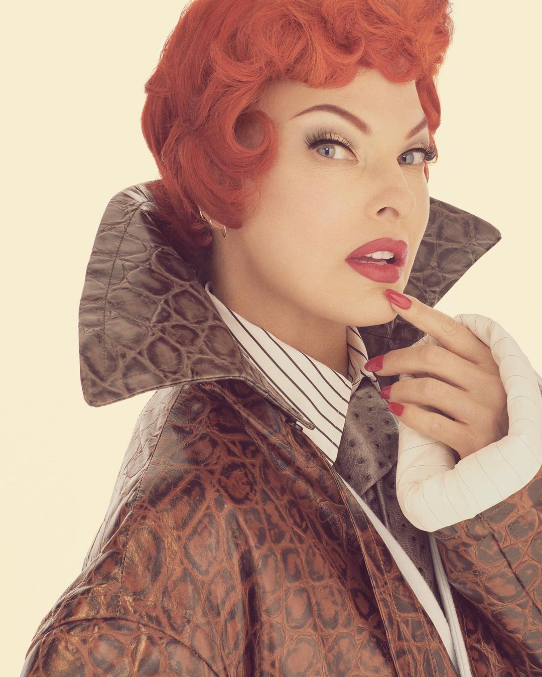Close-up of an adult woman with red curly hair, wearing a patterned jacket and striped shirt, looking at the camera in a photo studio.