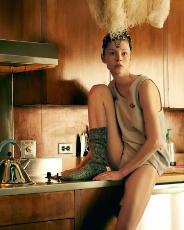 Stylish woman with feathered headpiece poses in retro-style kitchen with kettle, stove, and wood paneling in a fashion editorial shot.