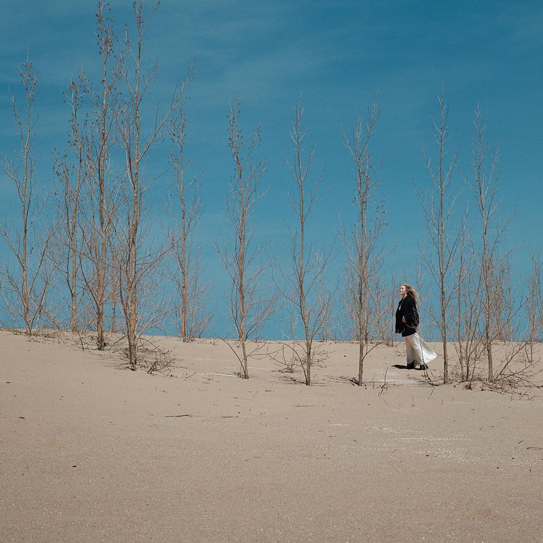 Woman in dark coat and flowing white skirt standing in a sandy desert landscape among bare trees under a clear blue sky