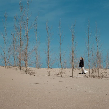 Woman in dark coat and flowing white skirt standing in a sandy desert landscape among bare trees under a clear blue sky