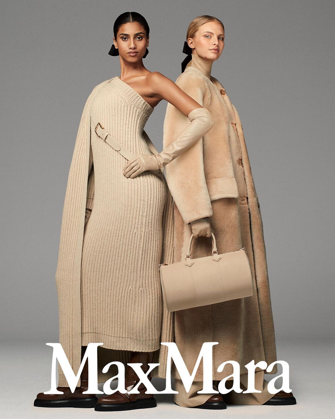 Two women model neutral-toned Max Mara knitwear, a wool coat, and leather accessories in a studio shot with a muted gray background.