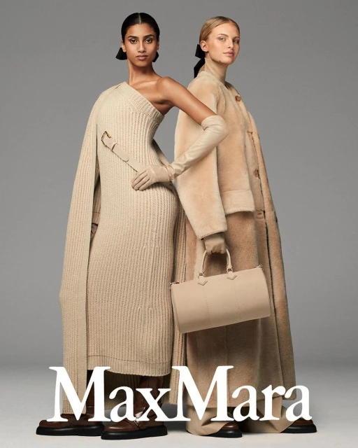 Two women model neutral-toned Max Mara knitwear, a wool coat, and leather accessories in a studio shot with a muted gray background.