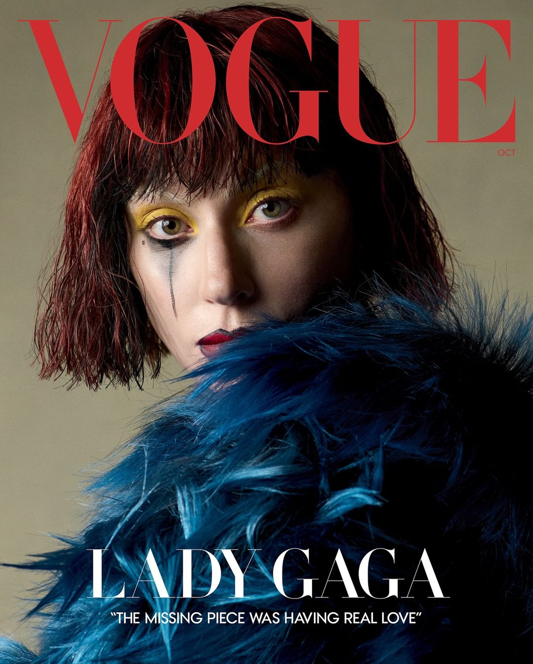 Vogue cover featuring Lady Gaga with dramatic yellow eye makeup, dark lip, and teal feathered boa. Dark red hair and neutral background.