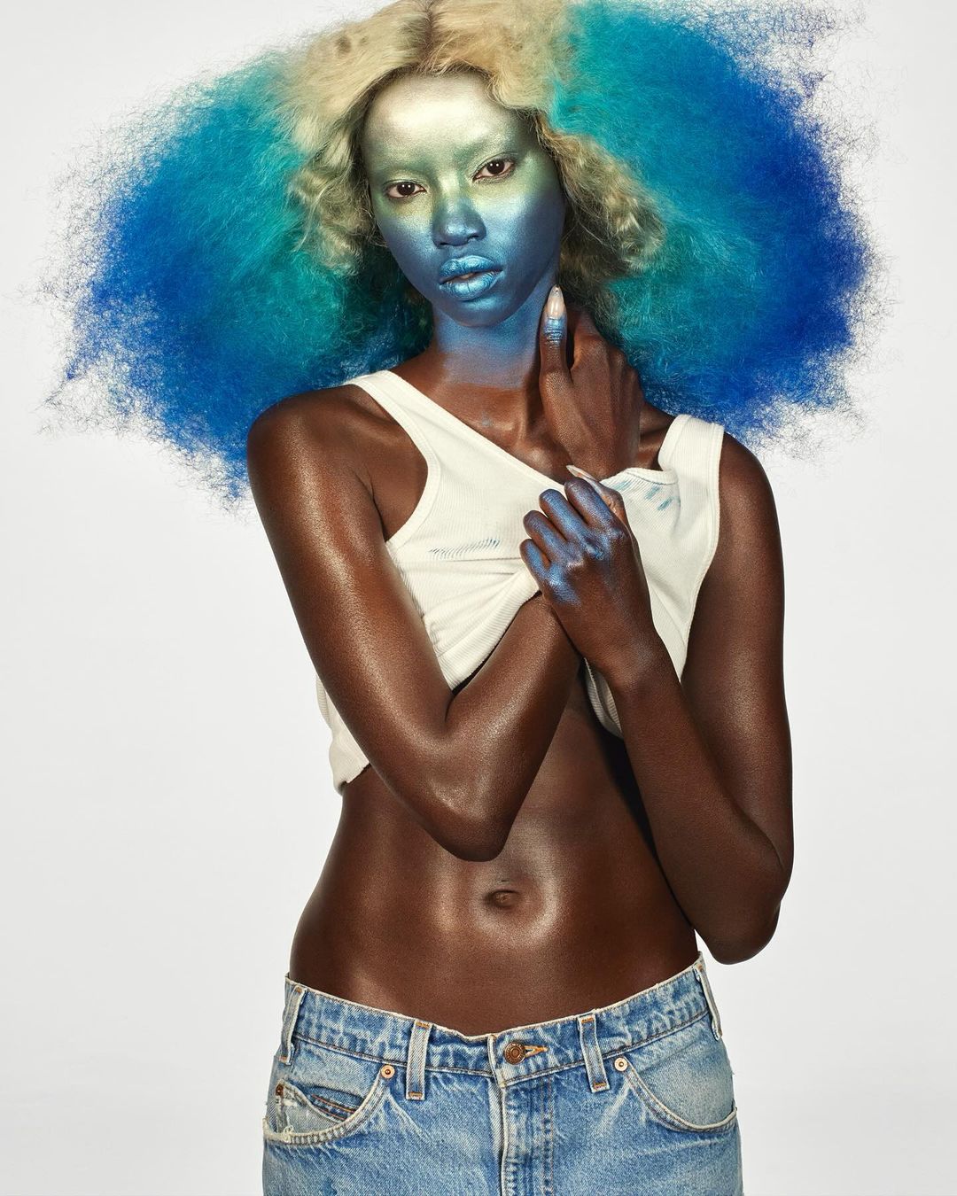 Stunning studio shot of a Black female model with unique blue and blonde hair, eye-catching makeup, tank top, and jeans against a white backdrop.