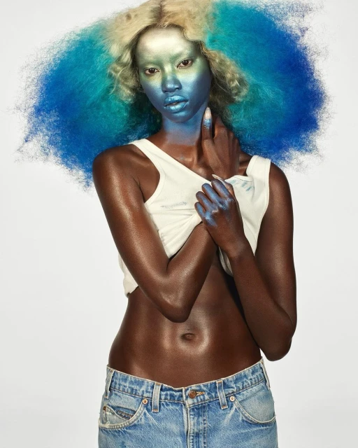 Stunning studio shot of a Black female model with unique blue and blonde hair, eye-catching makeup, tank top, and jeans against a white backdrop.
