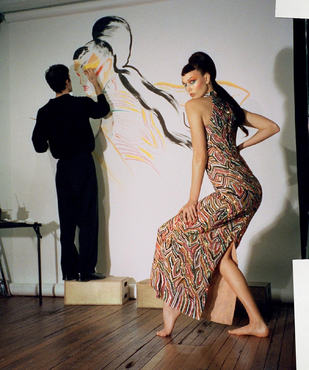 Fashion shoot features a model in patterned dress posing next to an artist painting a portrait on a large canvas in a studio space.