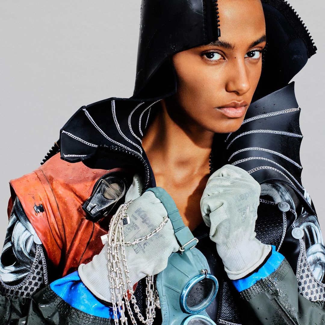 Portrait of an adult female fashion model wearing a black hooded jacket and holding a gas mask, standing in front of a solid grey background.